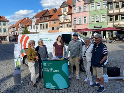 Petra (dritte von links) steht mit  fünf Senioren, darunter Mitglieder des Seniorenbeirats, an dem Info-Stand auf dem Marktplatz in Helmstedt. Im Hintergrund sind historische Häuser zu sehen.