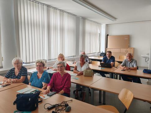 Fünf Damen und drei Herren sitzen an drei Tischen im Raum und blicken nach Vorne in die Kamera.