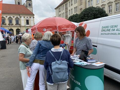 Mobilreferentin Theresa erklärt 4 Seniorinnen etwas am Infostand auf dem Marktplatz. Rechts neben ihr ist das Infomobil.