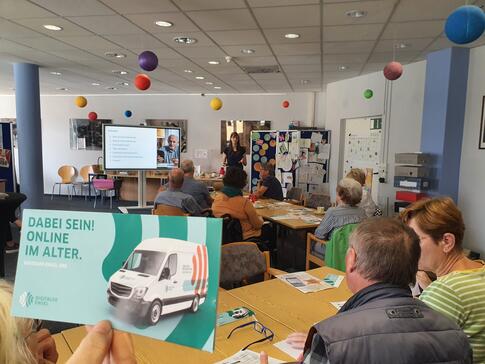 Links im Vordergrund ist ein Flyer vom Digitalen Engel in der Luft gehalten. Rechts davon und im Hintergrund sitzen ältere Personen an Tischen. Ganz weit im Hintergrund steht Petra neben einer digitalen Tafel.