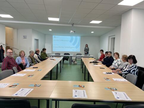 Die 12 älteren Teilnehmenden sitzen an Tischen in einer U-Form und lächeln in die Kamera. Theresa steht rechts neben der Leinwand und lächelt ebenso.