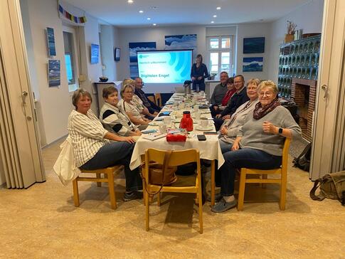 An einem langen Tisch sitzen zwölf Senior:innen (die meisten davon Frauen) und blicken nach vorne in die Kamera. Im Hintergrund steht Petra mit einem großen Bildschirm.