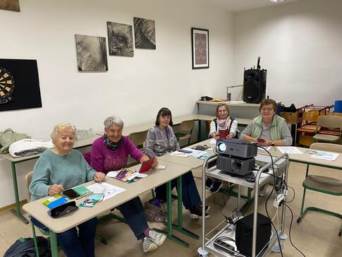 Fünf ältere Damen sitzen an einer kurzen Tischreihe und lächeln in die Kamera. Vor ihnen steht auf einem Podest ein Beamer.