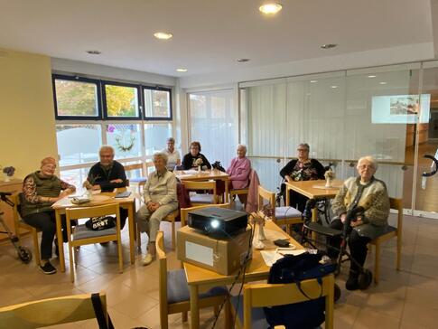 Sieben Damen und ein Mann sitzen in einem Raum verteilt an drei Tischen. Im Vordergrund steht ein Beamer. Im Raum stehen zwei Rollatoren.