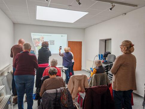 Eine Gruppe von Menschen steht in und sitzt einem Raum und mehrere Personen halten ihr Handy hoch um einen QR-Code einzulesen, der auf eine Wand projjiziert ist