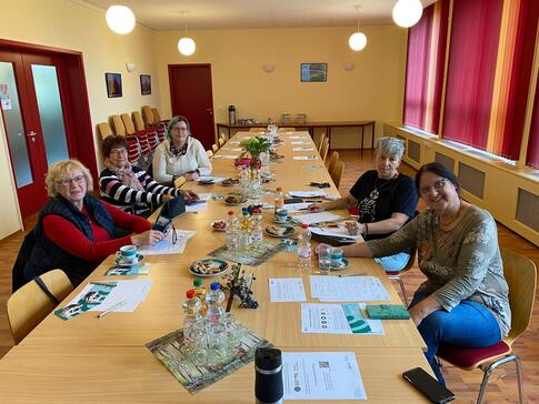 Fünf Frauen sitzen an einem Tisch gegenüber und schauen in die Kamera nach vorne. Auf dem Tisch liegen Materialien und Herbstdeko.