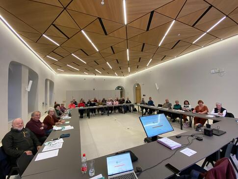 Auf dem Bild im Weitwinkelmodus sitzen in einem großen Sitzungssaal ältere Menschen an Tischen, die in U-Form aufgebaut sind. Im Innenraum der Tische steht ein Bildschirm.