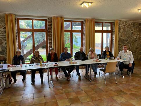 An einem langen Tisch sitzen acht Personen (drei M&auml;nner und f&uuml;nf Frauen) und gucken nach vorne in die Kamera.