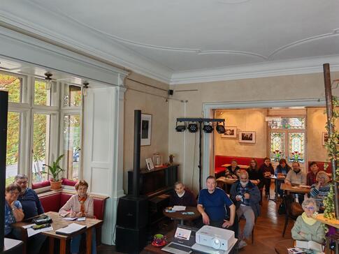 Mehrere Personen sitzen an kleinen Tischen im Raum und blicken nach Vorne. 