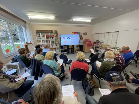 Die Teilnehmenden sitzen auf Stühlen in Halbkreisen und sind von hinten zu sehen. Vorne steht Mobilreferentin Theresa rechts neben der Leinwand und lächelt in die Kamera.