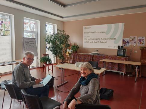 Ein Herr und eine Dame sitzen auf Stühlen und drehen sich und blicken in Richtung Kamera. 