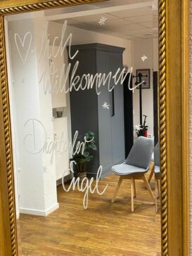 Auf dem Spiegel steht in Schreibschrift der Text "Herzlich Willkommen Digitaler Engel". In dem Spiegel ist ein Schrank und ein Stuhl zu sehen.