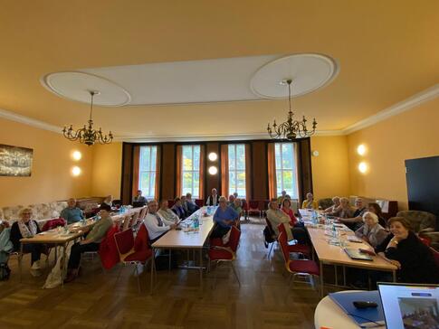 In einem großen Saal sitzen an drei langen Tischen insgesamt knapp 30 Senior:innen.