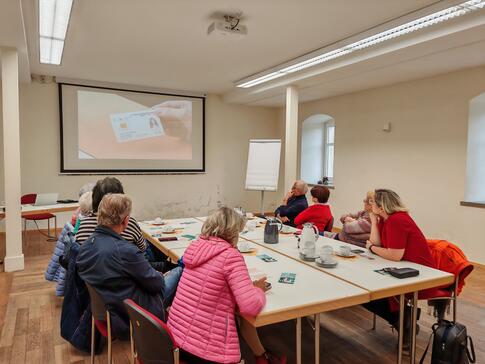 Mehrere Personen sitzen um einen zusammengeschobenen Tisch und blicken auf eine Leinwand, auf der ein Video gezeigt wird.