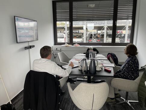 Zwei Teilnehmende sitzen in einem kleineren Besprechungsraum an einem Tisch. Sie sind von hinten oder von der Seite zu sehen. Links im Raum ist der Monitor mit der ersten Folie der Präsentation. Im Hintergrund ist ein Fenster, durch das Autos zu sehen sind.