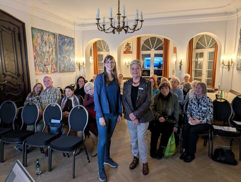 Petra (links) steht neben einer Ansprechpartnerin der Loge vor dem Publikum, welches auf mehreren Stuhlreihen sitzt.