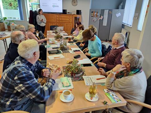 Die Teilnehmenden sitzen an einer langen Tischreihe und schauen zu Theresa, die das Projekt Digitaler Engel vorstellt.