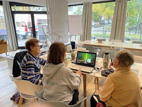 Theresa und zwei Teilnehmerinnen sitzen an einem Tisch und sind von hinten zu sehen. Theresa zeigt den beiden älteren Frauen Dinge am Laptop.