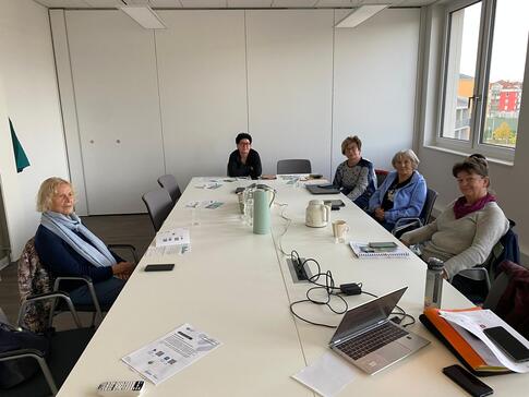 In einem Besprechungsraum sitzen an einem rechteckigen Besprechungstisch vier Seniorinnen. Auf dem Tisch liegen Materialien und ein Laptop.