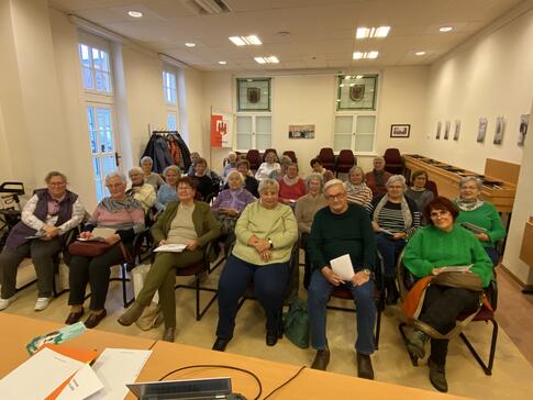 In einem gelb erleuchteten Raum sitzen in vier Sitzreihen 25 Senior:innen. Sie blicken nach vorne in die Kamera.