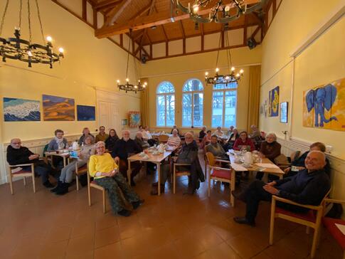 Im B&uuml;rgertreffpunkt im Bahnhof Lichterfelde sitzen an verschiedenen Gruppentischen 27 Senior:innen und blicken nach vrone in die Kamera.