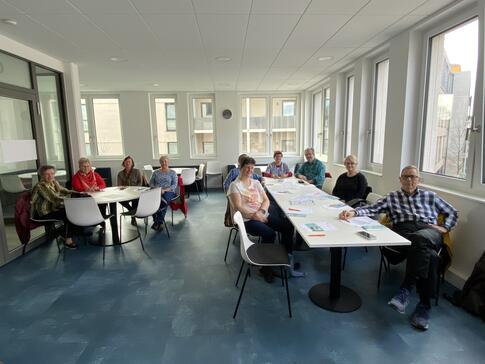 In einem Raum sitzen an einem langen Tisch und an einem runden Tisch insgesamt 11 Senior:innen und blicken nach vorne in die Kamera.