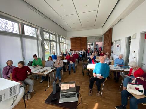 In einem gro&szlig;en Raum sitzen &uuml;ber drei&szlig;ig Senior:innen an mehreren Tischen frontal nach vorne und blicken in die Kamera.