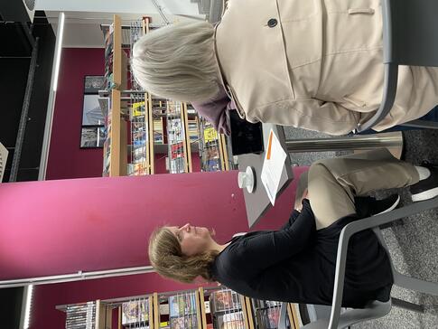 Mobilreferentin Theresa sitzt mit einer &auml;lteren Frau und dem Radiojournalisten an einem Tisch. Alle sind von hinten oder der Seite zu sehen. Im Hintergrund sind B&uuml;cherregale.