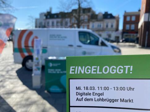Im Hintergrund steht das Info-Mobil bei strahlendem Sonnenschein mit dem Info-Stand. Im Vordergrund ist ein Flyer mit der Aufschrift "eingeloggt!". Darunter steht "Mi, 18.03. 11-13 Uhr. Digitale Engel. Auf dem Lohbr&uuml;gger Markt"