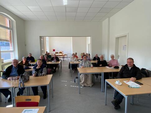 In einem Schulraum sitzen knapp 17 Senior:innen an Schultischen, auf denen Materialien liegen.