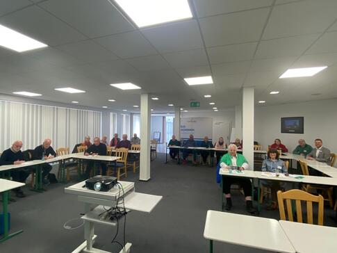 In einem gro&szlig;en Raum mit zwei Pfeilern in der Mitte sitzen Menschen an Tischen in U-Form. Im Vordergrund steht ein Beamer.