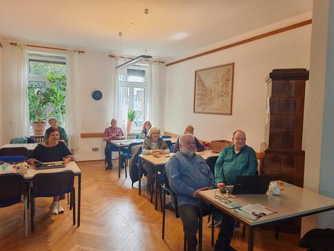 Mehrere Personen sitzen an Tischen verteilt im Raum und blicken nach Vorne.