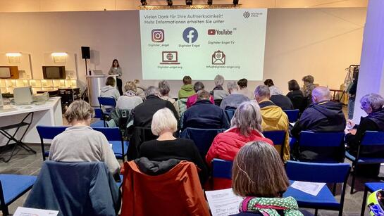 Die Teilnehmenden sitzen auf St&uuml;hlen im Computermuseum und sind von hinten zu sehen. Theresa steht zwischen Leinwand und alten Computern und l&auml;chelt in die Kamera.