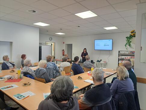 Theresa steht links neben einem Fernseher und stellt den Digitalen Engel. Die Teilnehmenden schauen zu ihr und sind von hinten oder der Seite zu sehen.