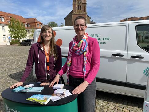 Mobilreferentin Petra (links) steht mit Ehrenamtskoordinatorin Danica Jansen (rechts) vor dem Info-Mobil am Info-Stand. Beide posieren und l&auml;cheln in die Kamera. 