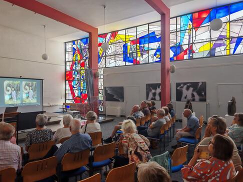 Zahlreiche Personen sitzen auf St&uuml;hlen im Raum verteilt und blicken auf eine Leinwand auf der ein Video gezeigt wird.