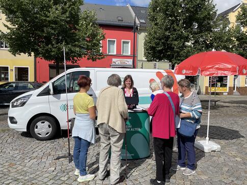 Mobilreferentin Theresa zeigt 5 &auml;lteren Frauen etwas auf dem Tablet. Sie stehen am Infostand auf einem Marktplatz. 