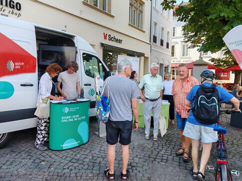 Mehrere Personen stehen auf einem Platz vor dem Infomobil, eine Dame l&auml;sst sich von Referent Johannes etwas auf ihrem Tablet zeigen.