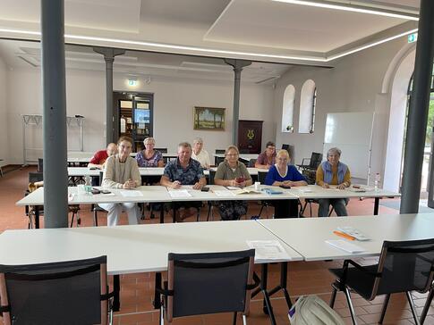 Die Teilnehmenden sitzen in zwei Tischreihen hintereinander und l&auml;cheln in Kamera.