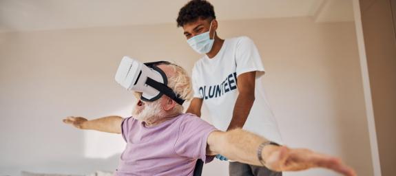 Älterer Herr im Rollstuhl trägt eine VR Brille und wird von einem jungen lächelnden Mann geschoben.