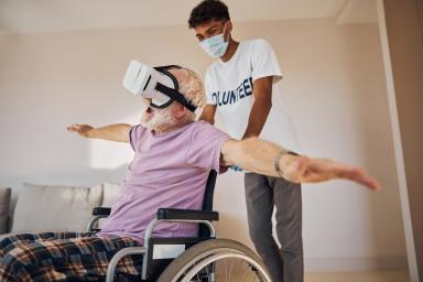 junger Mann schiebt Älteren im Rollstuhl mit ausgebreiteten Armen und VR Brille